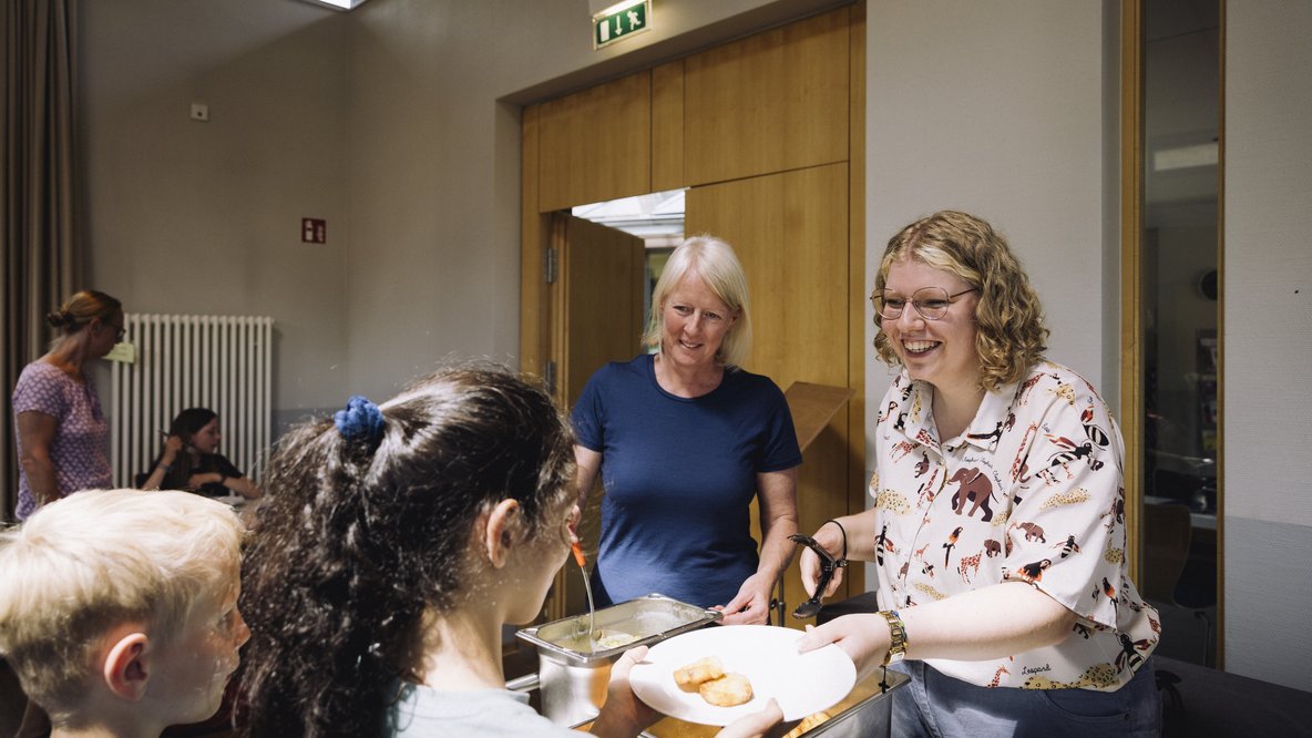 Zwei Frauen lächeln Kinder an und eine Frau verteilt Essen auf einem Teller an ein Kind.